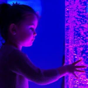 A young girl in a sensory room reaching out to touch a free-standing bubble tube - representing the Purple House Clinic's sensory assessment service.