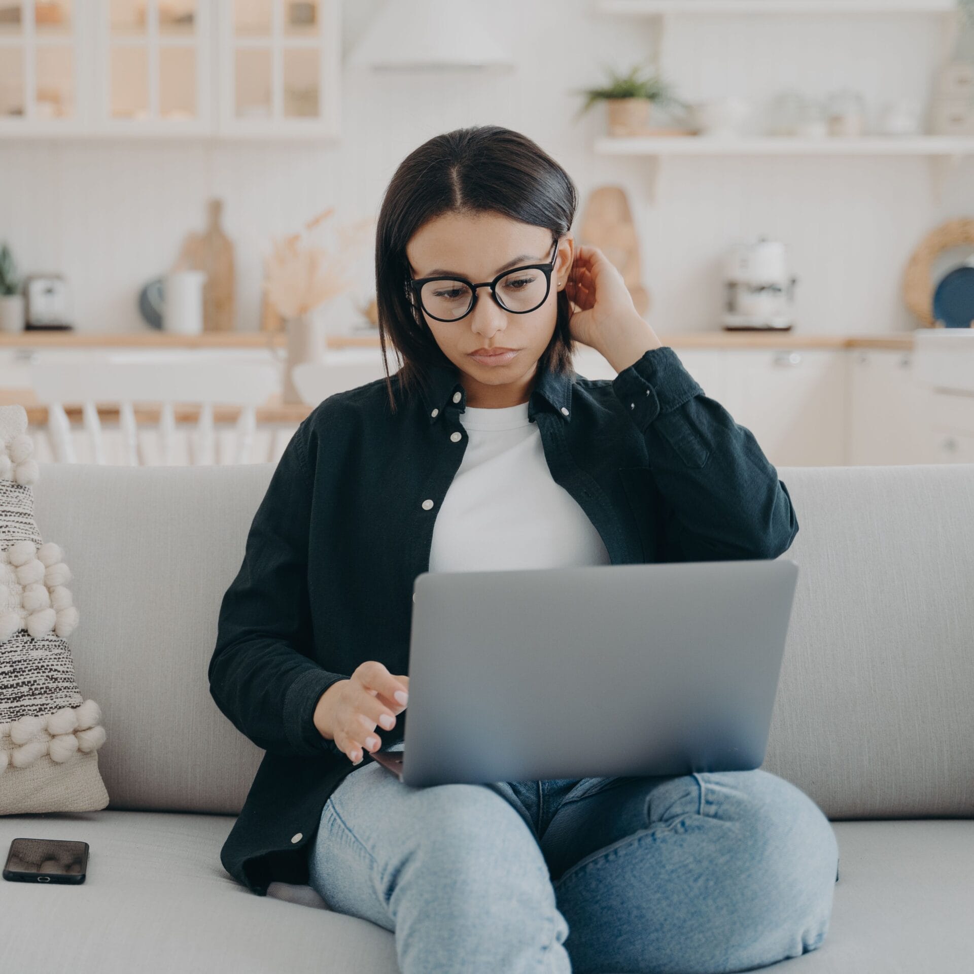 A young woman wearing glasses sits cross-legged on sofa at home during an online therapy session